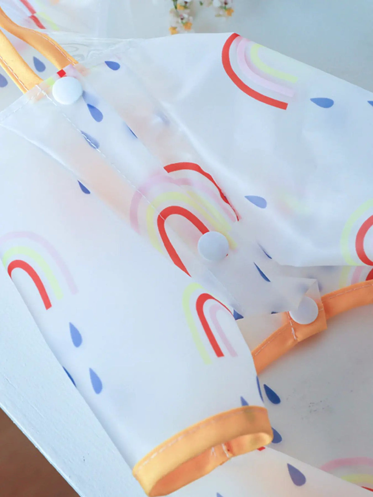 Lightweight waterproof pet raincoat, white with pastel rainbow arches, blue droplets, and orange trim with snap closures