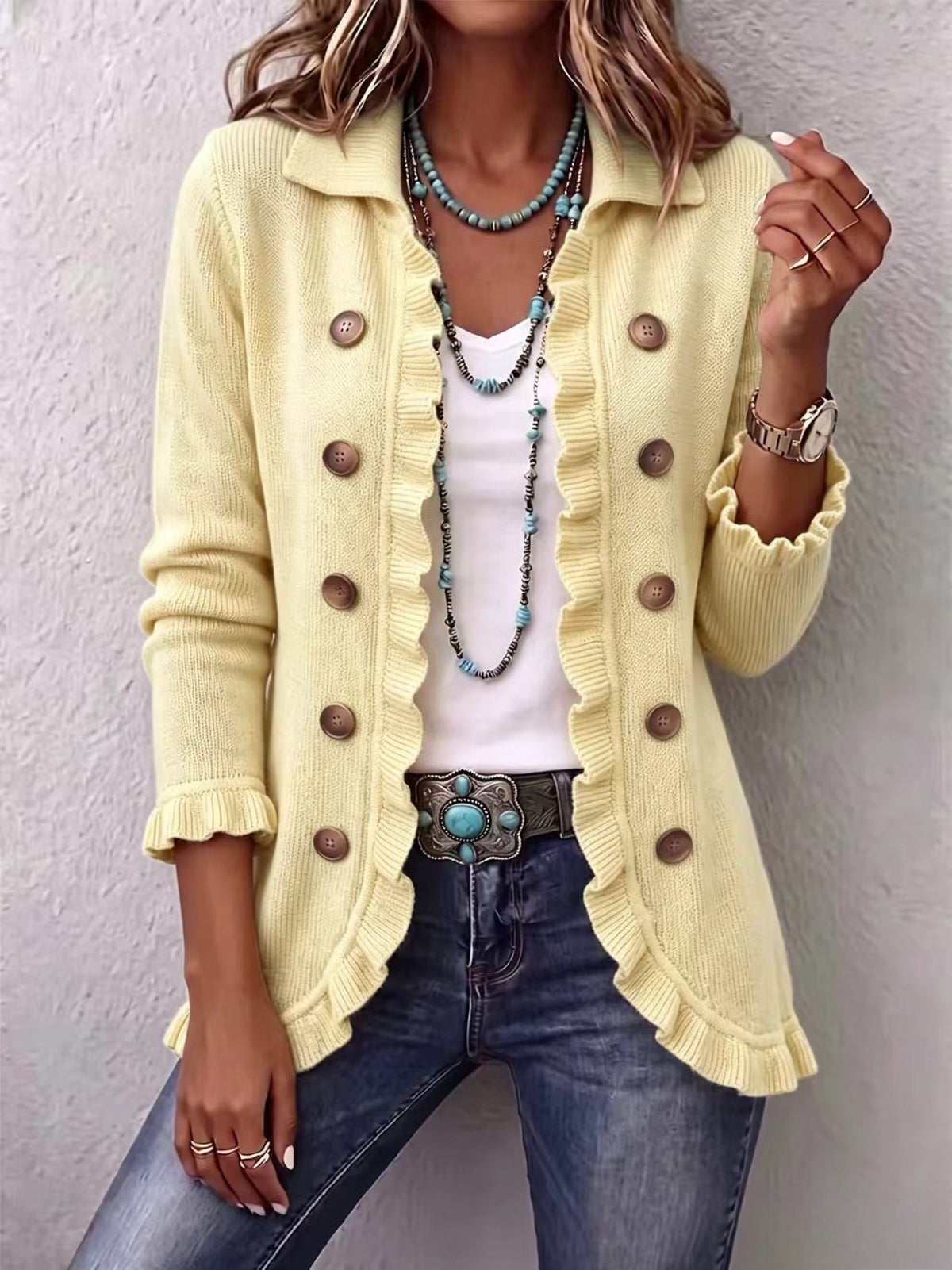 Long pale-yellow knit cardigan with ruffled edges and large brown buttons, layered over a white top.