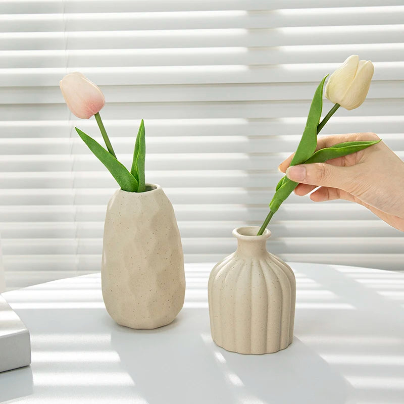 Ceramic vase pair in beige with ribbed and speckled finishes, pale tulips on a white table; hand placing a stem.