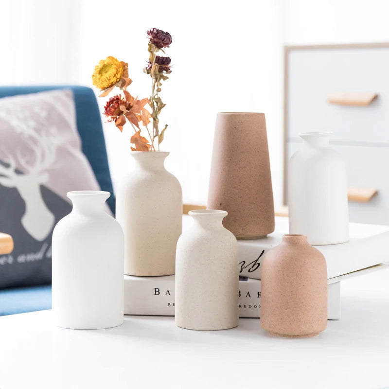Ceramic vase set in white, cream, tan and terracotta tones; assorted shapes on a white table, one with dried flowers.