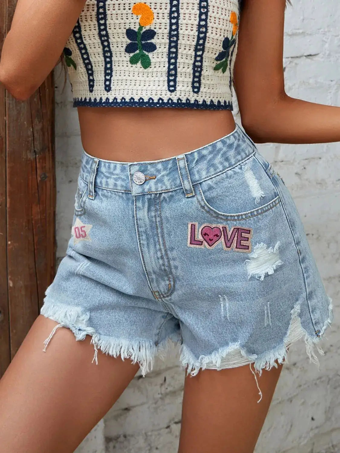 Women's light-wash denim shorts with distressed raw-hem, button front, four pockets, and pink LOVE patch.