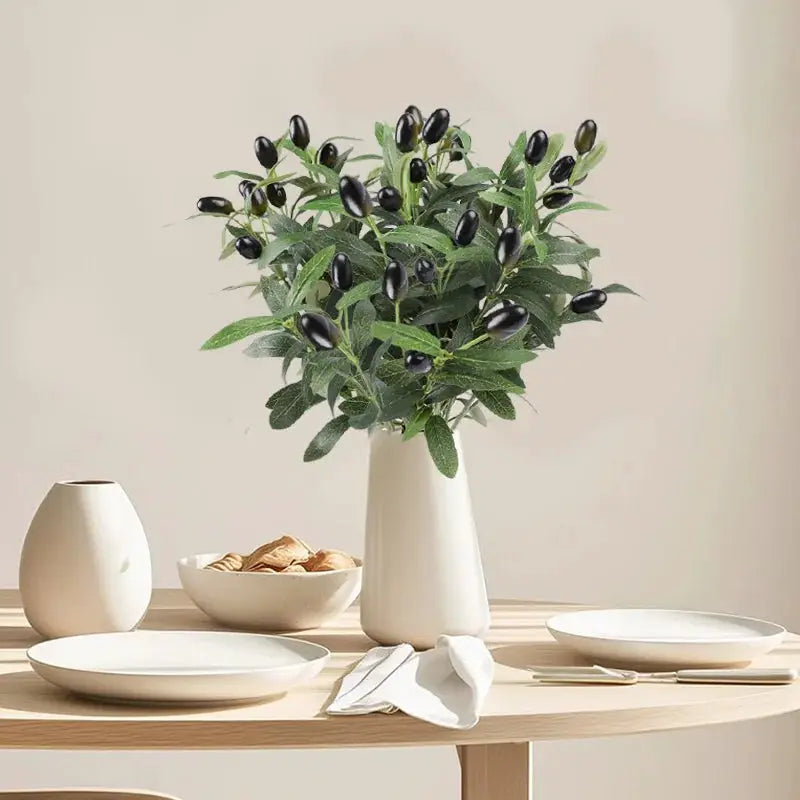 Artificial olive branch with green leaves and black olives in a white vase on a light wood table.