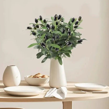 Artificial olive branch with green leaves and black olives in a white vase on a light wood table.