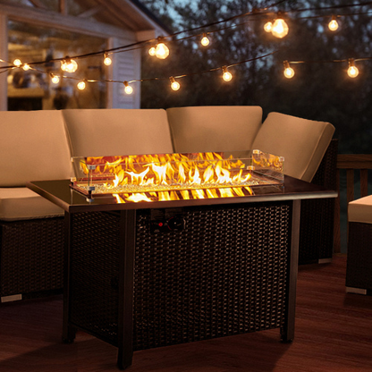 Rattan propane outdoor fire pit table with glass wind guard; dark wicker on a wooden deck with string lights.